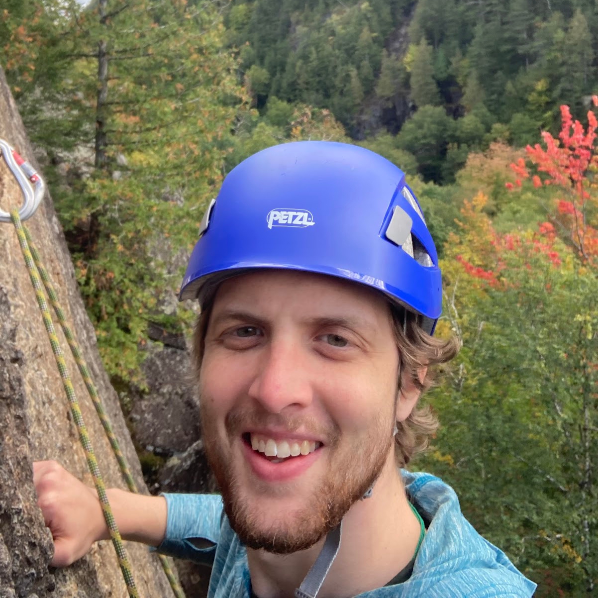 a young man wearing a blue helmet faces the camera as he climbs a sheer rock face, he is smiling in front of rolling hills of fall leaves