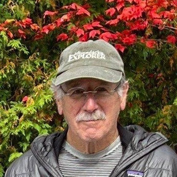 An older man wearing a hat stands in front of a wilderness scene with green leaves and red flowers